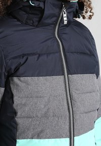 Enfant portant une doudoune color-block avec des panneaux noirs, gris et bleu sarcelle, et une fermeture éclair à l'avant remontée jusqu'au cou.