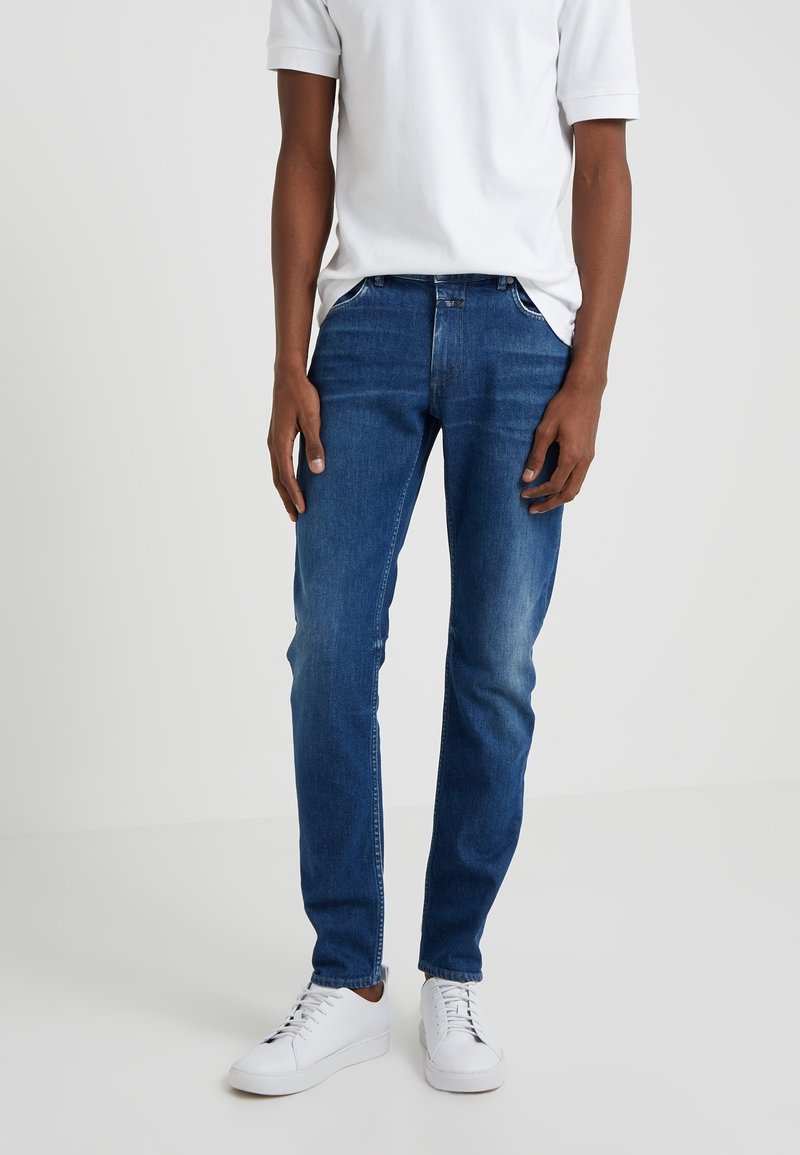 Homme portant des baskets blanches, un jean bleu slim et un t-shirt blanc uni à manches courtes, debout devant un fond clair.