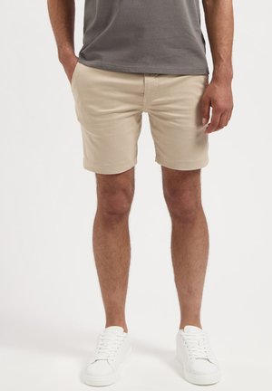 Man wearing beige shorts, gray shirt, white sneakers, standing with one hand in pocket against white background.