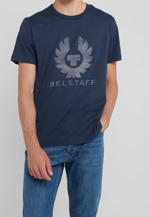 Homme portant un t-shirt Belstaff bleu marine avec logo et texte, associé à un jean bleu, main dans la poche, sur fond gris uni.