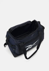 Sac de sport noir avec un extérieur texturé, fermeture éclair, doubles poignées et un logo argenté proéminent d'un côté. Intérieur ouvert visible.