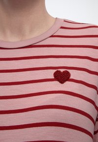 Κοντινό πλάνο ριγέ πουκάμισου σε ροζ και κόκκινους τόνους με ένα μικρό κόκκινο υφασμάτινο καρδιά στο αριστερό μέρος του στήθους.