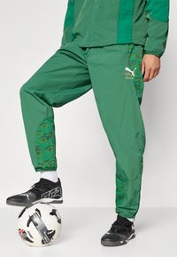 Zelene trenirke s prostim krojem, ki imajo camo vzorec na spodnjih nogavicah in izvezeno logo. V paru z črnimi športnimi čevlji na nogometni žogi.