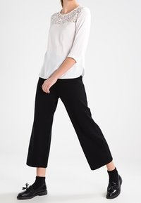 Vit blus med spetsdetaljer vid halsringningen och 3/4-långa ärmar, i kombination med svarta vida byxor och svarta snörskor.