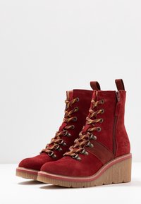 Botas de tobillo de ante rojo con detalles en encaje dorado, cremallera lateral y suela de plataforma de goma. Presenta una correa de cuero suave alrededor del tobillo.