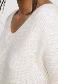 Biały sweter z dzianiny o dekolcie w kształcie V i prążkowanej strukturze, prezentujący miękki i lekki materiał.