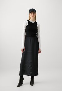 Svart ermeløs topp brukt over et langt, svart perforert skjørt, kombinert med en svart caps og spisse svarte høyhælte støvler.