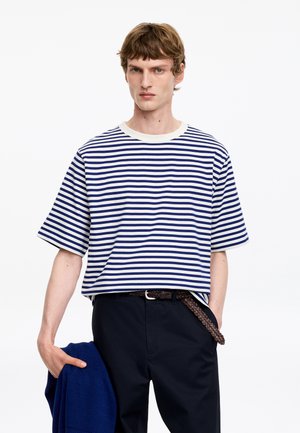 Homme portant une chemise à manches courtes rayée blanche et bleu marine, pantalon noir avec ceinture tressée, tenant un pull bleu dans la main gauche sur fond blanc.