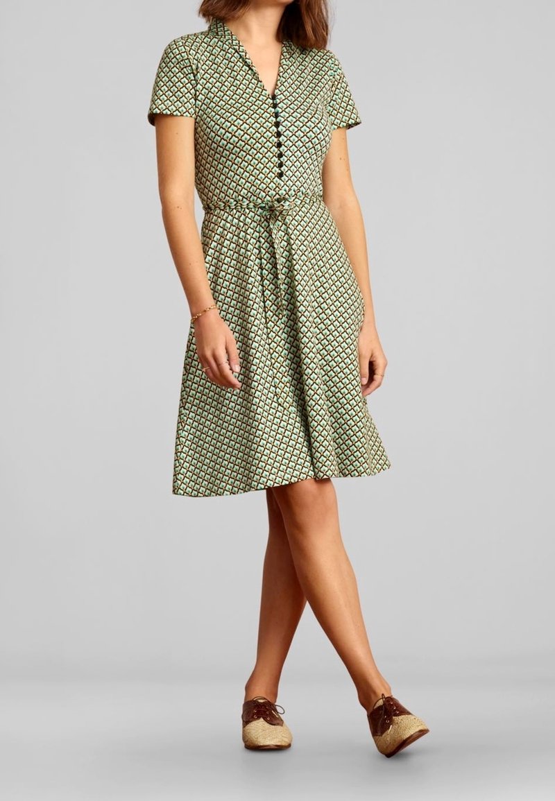 Zielona sukienka w wzory z krótkimi rękawami, zapinana na guziki, z wiązanym w talii detalem; zestawiona z brązowymi butami na sznurówki.
