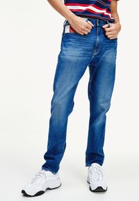 Hombre vestido con jeans azules y zapatillas blancas, posando con las manos en la cintura, camiseta a rayas rojas y azules visible en la parte superior.