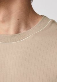 Beige, ribbet striktrøje med en kontrasterende, lyse farvet halsudskæring. Glat tekstur, tætsiddende pasform og subtile detaljer langs kanterne.