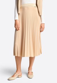 Falda midi plisada en beige con cinturilla texturizada. Combinada con zapatos de tacón bajo tipo slingback en beige con dobles tiras.