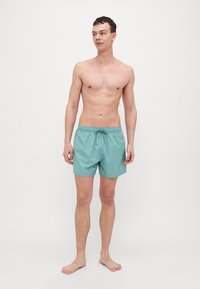 Vaalean teal-väriset uimapuvut, joissa on joustava vyötärö ja nyöritys, valmistettu sileästä ja kevyestä materiaalista, jossa on huomaamaton logo.