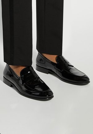 Black patent leather loafers with a smooth finish, featuring a decorative strap and low block heel, worn with tailored black trousers.