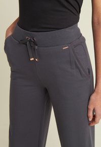 Pantalones de chándal grises con una textura suave, cinturilla elástica, cordón y bolsillos laterales. Pequeños acentos metálicos añaden detalle.