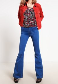 Červená semišová bunda přes květinové tričko, kombinovaná s rozšířenými modrými džíny a černými botami na podpatku. Outfit se vyznačuje živými barvami a různými texturami.
