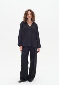 Blusa nera, ampia e a maniche lunghe con scollatura a V e due tasche davanti, abbinata a pantaloni neri a gamba larga e scarpe nere a punta.