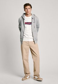 Sudadera gris con cremallera sobre una camiseta blanca con un logo rojo y azul, combinada con pantalones beige y zapatillas con acentos marrones.