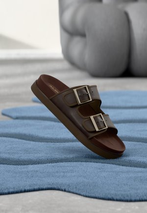 Sandale à glissière en cuir marron avec deux boucles argentées réglables, posée sur une surface texturée bleue, fond neutre.