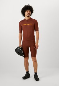 Bruine fietsshirt en shorts met een aansluitend ontwerp en korte mouwen. Voorzien van een rits aan de voorkant en een 'Castelli' logo in geel. Zwarte fietsschoenen in hand.