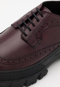 Chaussure brogue en cuir bordeaux foncé avec des lacets ronds noirs et une semelle en caoutchouc noire épaisse et texturée, affichant un design perforé détaillé.