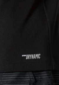Černé bavlněné tričko s hladkou texturou, na kterém je v bílé barvě nápis "DRYKORN DRYNAMIC". Design zahrnuje zakřivený lem a jemné švy.