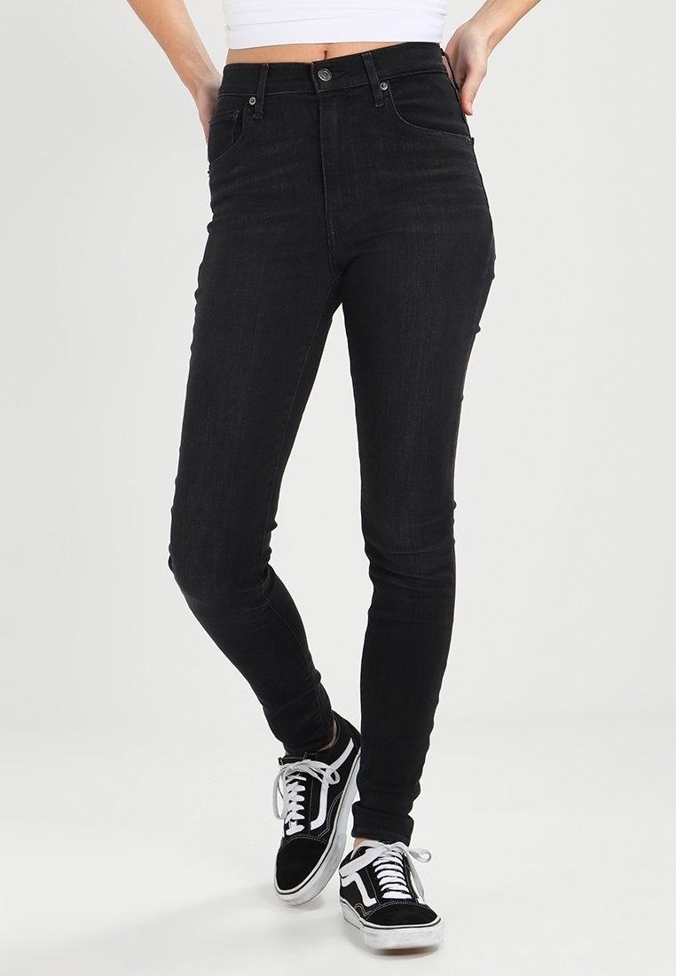 Jeans ajustados de tiro alto en color negro, confeccionados en denim, con bolsillos frontales, cremallera y cierre de botón. Combinados con zapatillas negras y blancas.