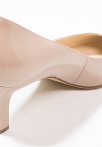 Chaussure à talon haut beige avec une finition vernie brillante, bout arrondi et silhouette épurée. Détail texturé sur le côté et talon étroit.