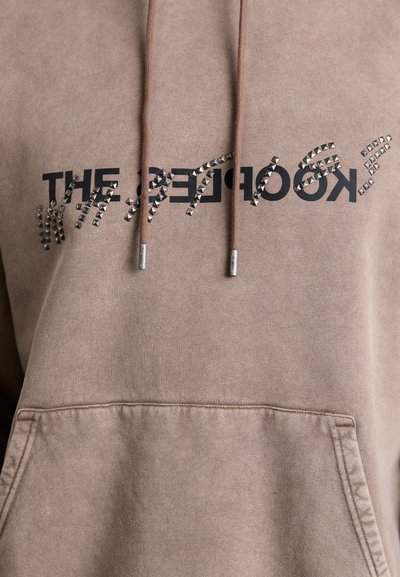 Brauner Hoodie aus weichem Stoff mit einer vorderen Tasche, Kordelzügen und einem schwarzen bedruckten Logo, verziert mit silbernen Nieten.