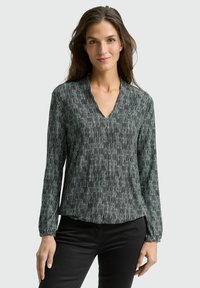 Woman wearing a long-sleeve black and white patterned V-neck blouse and black pants, standing against a light gray background.