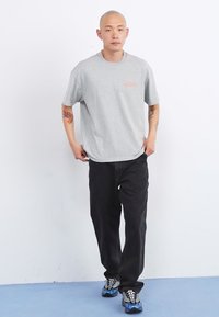Gray cotton t-shirt with a loose fit and orange logo, paired with black jeans and multicolored athletic shoes. Plain background.