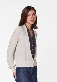 Lichtgrijze cardigan met een textuurdesign, open voorkant, geribde manchetten en een knoopsluiting bij de taille, gedragen over een donkere top en denim.
