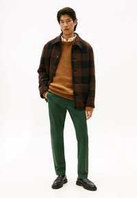 Brązowy i czarny w kratę płaszcz na pomarańczowym teksturowanym swetrze, w połączeniu z zielonymi spodniami i czarnymi butami. Prosty wzór, luźny krój.