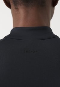 Čierne športové tričko s jemnou textúrou. Vysoký golier a logo "STRONGER" s vystúpeným písmom na chrbte. Jednoduchý dizajn, priliehavý strih.