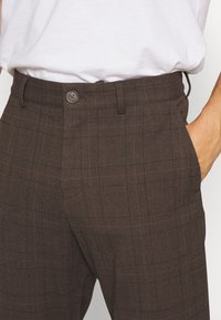 Bruna rutiga byxor med knappstängning, sidofickor och ett texturerat tyg, med en skräddarsydd passform och ett synligt midjeband.