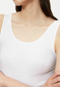 Woman wearing a white sleeveless tank top, arms crossed, with long brown hair and visible collarbone area on a plain background.