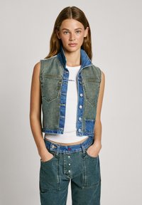 Džínová vesta s svetlomodrým a tmavomodrým farebným schémou, zapínanie na gombíky, dvoma náprsními vreckami a vysokým golierom. Nosená cez biely top.