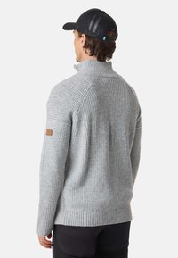 Man wearing light grey ribbed sweater and black cap, facing away, against plain white background.