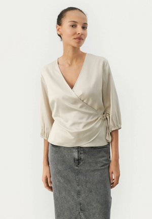 Beige wrap blouse with a deep v-neck and three-quarter sleeves, featuring a gathered side accent. Paired with a gray denim skirt.