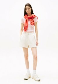 Shorts blancos y una camiseta ajustada rosa, con un sudadera roja atada alrededor de los hombros. El modelo lleva zapatillas blancas y calcetines cortos.