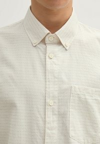 Knopfhemd aus hellbeigem Stoff mit kleinen, mehrfarbigen Punkten, klassischem Kragen und einer Brusttasche. Zwei sichtbare Knopfverschlüsse.