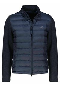 Navy-blue puffer vest with a zippered front and fleece sleeves. Horizontal quilted pattern on the torso; solid sleeves, logo on chest.