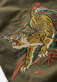 Vyšívaná nášivka zobrazujúca skákajúceho tigra v zlatej a čiernej farbe, obklopeného zeleným a koralovým lístím, na tmavozelenom olivovom pozadí.