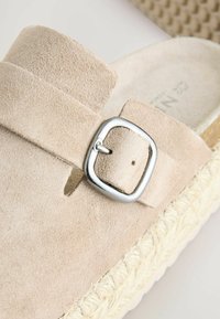 Sándalo de ante beige con una hebilla plateada, con una suela de yute trenzada estilo espadrille y una plantilla texturizada para mayor comodidad.