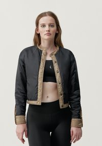 Svart kort bomberjacka med beige detaljer, tryckknappar och kviltad textur. Bärs över en svart sport-bh och åtsittande svarta leggings.