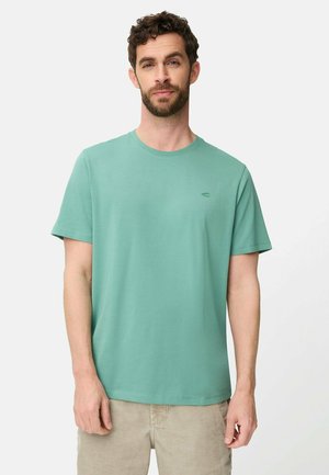 Mann mit kurzem, lockigem Haar und Bart, der ein einfaches, helltürkisfarbenes Kurzarm-T-Shirt und beige Hose trägt, vor einem weißen Hintergrund stehend.