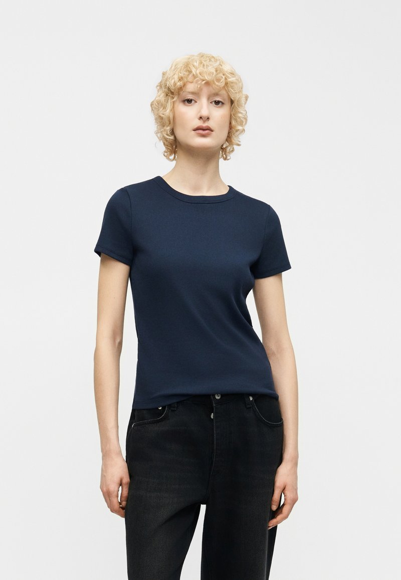 Personne aux cheveux blonds et bouclés, portant un t-shirt à manches courtes bleu marine et un jean noir, debout devant un fond clair uni.