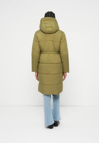 Casaco puffer verde-oliva com capuz, apresentando uma cintura ajustada, mangas longas e um design acolchoado até aos joelhos. Usado sobre uns jeans azul-claro.