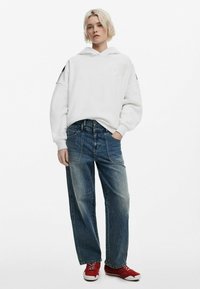 Hvid hoodie med en stor hætte og et subtilt logo, parret med blå brede denim jeans og røde sneakers.