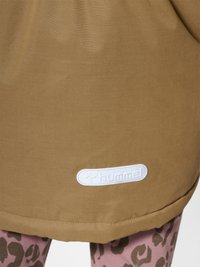 Spodní lem hnědé bundy s bílou nášivkou loga Hummel nad leopardími legínami na stojící osobě.